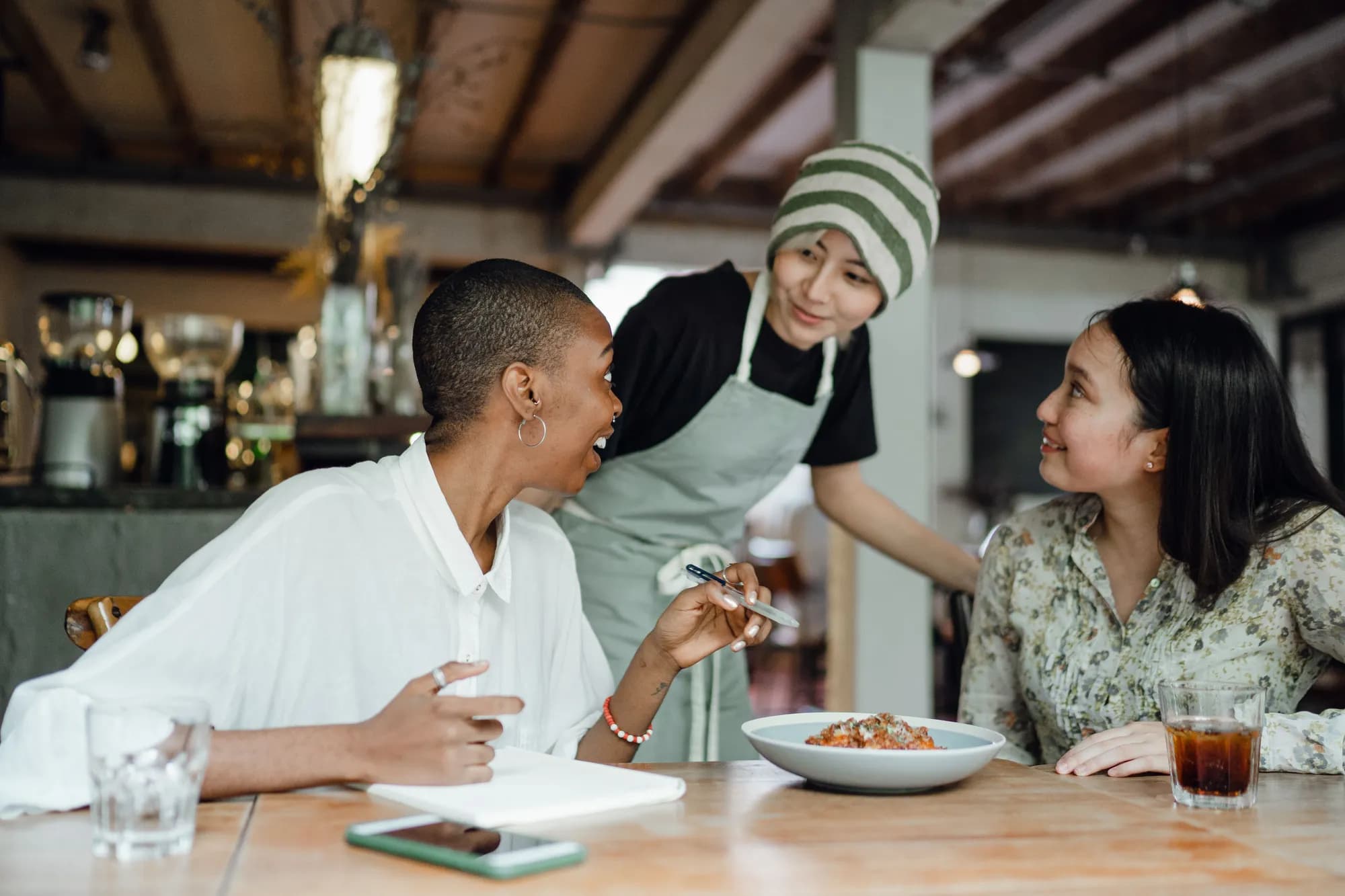 Atendente de touca anotando o pedido de duas pessoas à mesa de um café, entre risadas